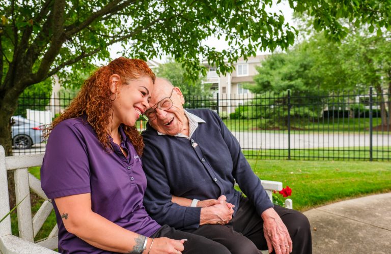 caregiver and assisted living resident sitting on bench smiling at each other and holding hands