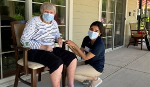 younger woman wearing mask kneeling next to an older woman wearing mask on a bench outside