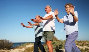 three older people with arms outstretched exercising on beach