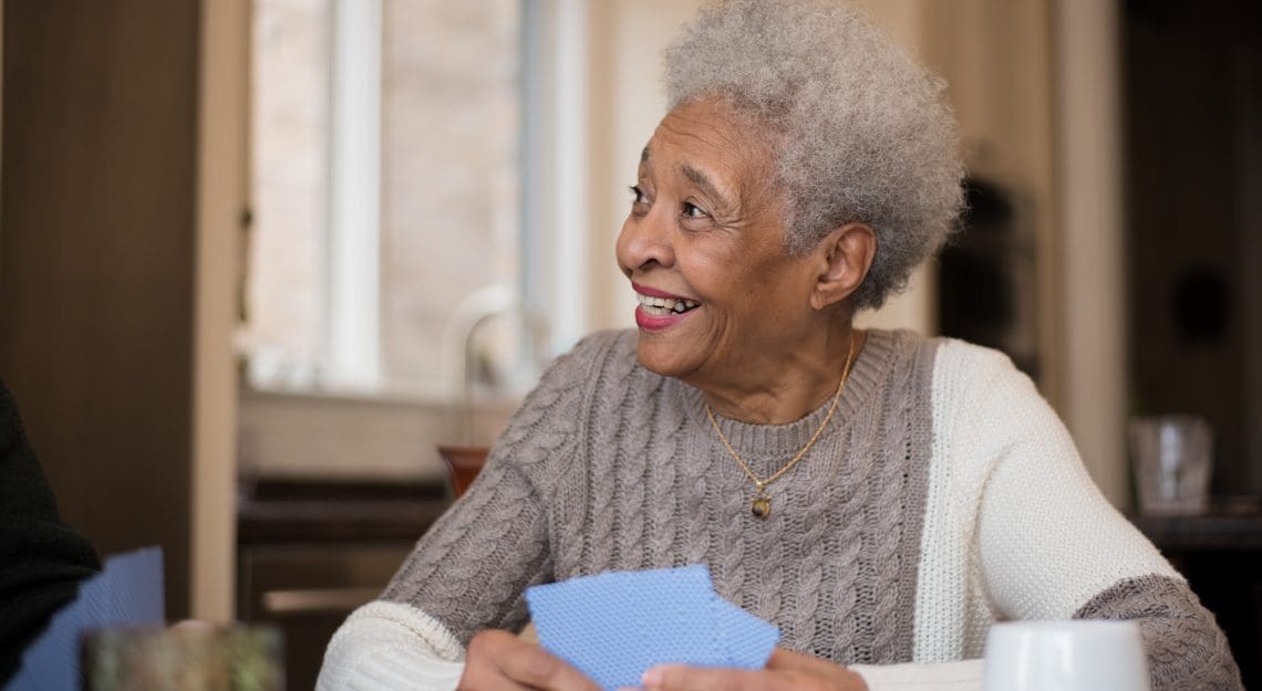 A senior woman of African descent smiles as she plays cards with friends off camera. She is also sitting at a kitchen table drinking coffee.