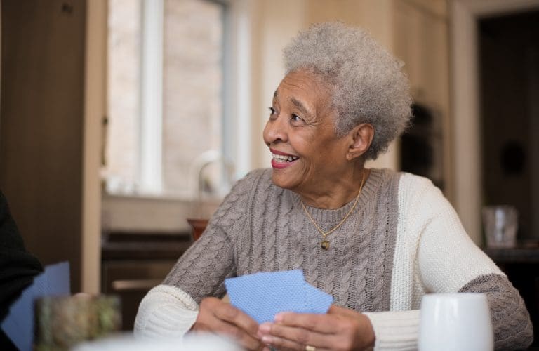 A senior woman of African descent smiles as she plays cards with friends off camera. She is also sitting at a kitchen table drinking coffee.