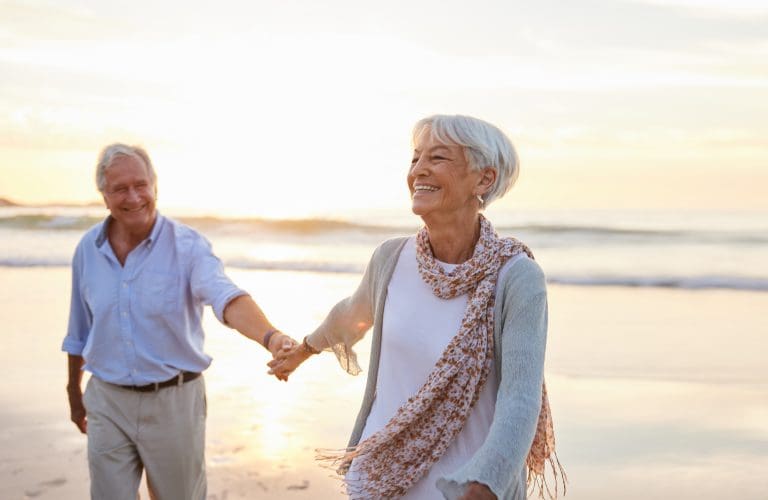 Senior woman laughing while leading her husband along a beach at sunset